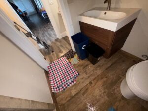 A high-angle shot of a bathroom floor showing signs of minor leaks, with water pooling on the wood-look tile near the toilet and sink. A red, white, and blue checkered towel and other damp cloths are placed on the floor to absorb the leaking water next to a blue recycling bin and a black trash can
