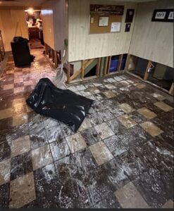 A basement room showing severe water damage and remediation efforts. The checkered tile floor is covered in a dirty, chalky white and brown water residue. Along the base of the wood-paneled walls, the lower section has been cut away and removed, exposing the internal wooden studs. A large black trash bag and a white plastic shovel rest in the center of the room.
