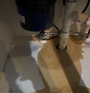 Under-sink cabinet displaying water damage around a PVC drain pipe, featuring peeling laminate and exposed, discolored composite wood.