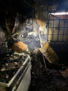 Severely fire-damaged kitchen corner with charred walls, soot-covered electrical appliances, and burnt debris.