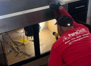 PuroClean of Southeast Tucson technician inspecting plumbing and water damage under a kitchen island sink.