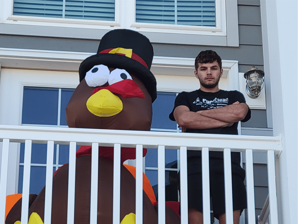 A young man stands on a porch next to a large inflatable turkey decoration for Thanksgiving.