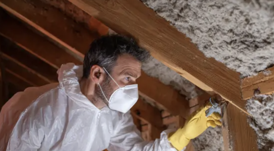 A man in a protective suit and mask applies sealant to attic insulation and wooden beams. TITLE