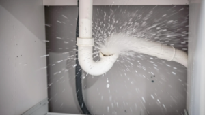 A white plastic drain pipe under a sink is actively spraying water from a significant leak.