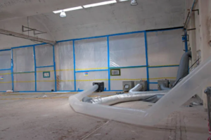 Large empty room with plastic-covered walls and flexible ventilation ducts on the concrete floor. TITLE