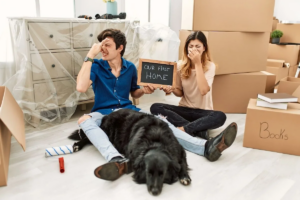 A couple and their dog pinch their noses in their new home, surrounded by moving boxes and holding