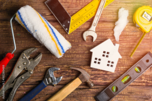 Various home improvement tools and a small house model are arranged on a rustic wooden surface
