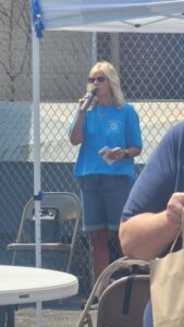 A blonde woman in sunglasses speaks into a microphone at an outdoor event under a white tent