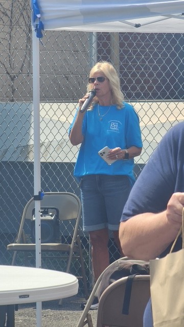 A blonde woman in sunglasses speaks into a microphone at an outdoor event under a white tent
