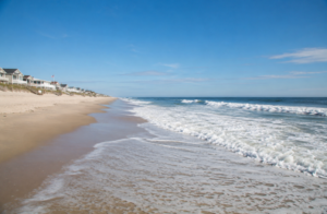 Beach houses line a sunny sandy shore with gentle ocean waves under a bright blue sky. TITLE