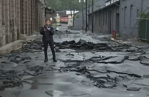 A reporter stands on a street with large sections of asphalt torn up by flood damage. TITLE