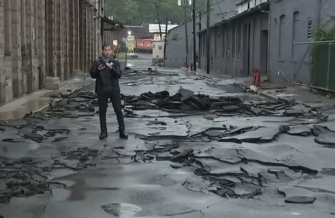 A reporter stands on a street with large sections of asphalt torn up by flood damage. TITLE