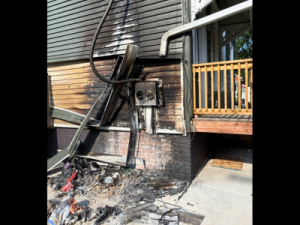Exterior wall of a house with heavy fire and smoke damage around the electrical panel, showing charring and soot that requires professional smoke damage restoration and safety repairs.