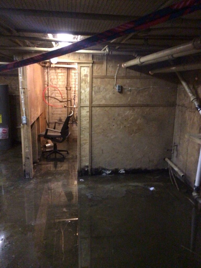 Flooded basement showing water accumulation on concrete floor with an office chair partially submerged, highlighting damage caused by minor leaks.