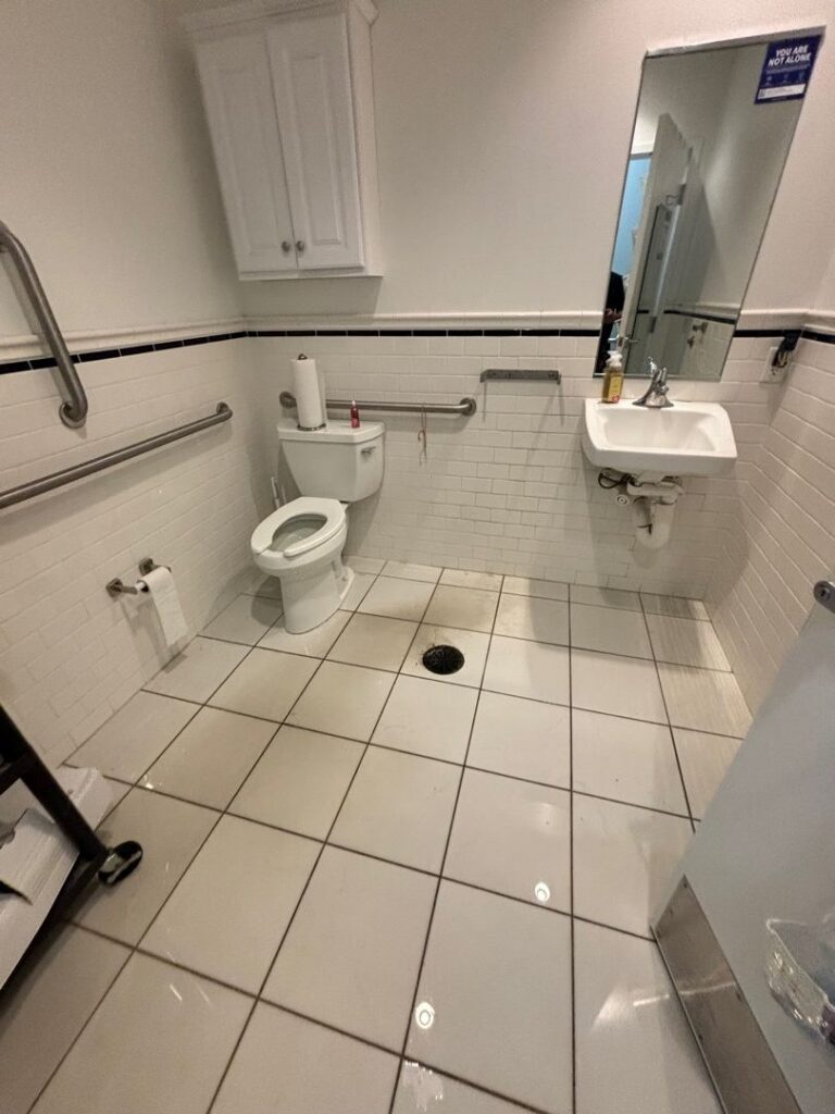 Bathroom with white tile flooring and accessible fixtures, showing standing water and wet floor conditions caused by a pipe burst, with water pooling around the drain, toilet, and sink area.