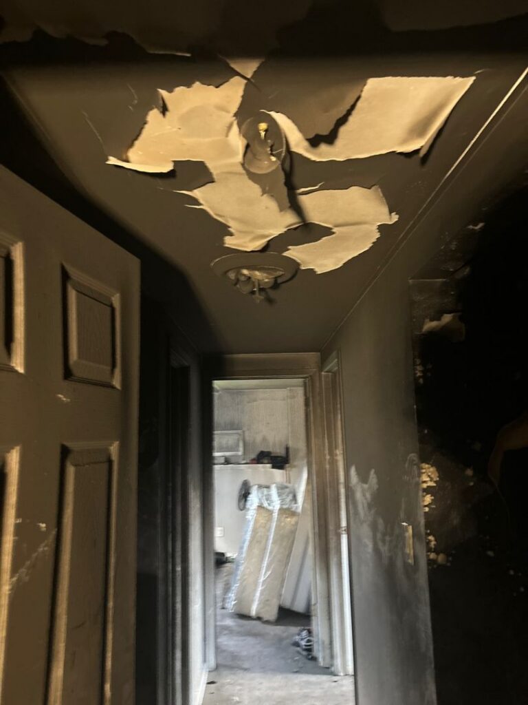 A narrow home hallway heavily damaged by fire, with the walls and ceiling covered in a thick layer of black soot, and sections of the ceiling paint peeling away to reveal lighter patches underneath.