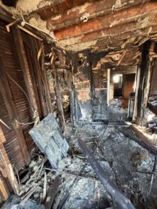 An interior view showing severe structural damage from a house fire, with exposed wooden wall studs and floor debris.