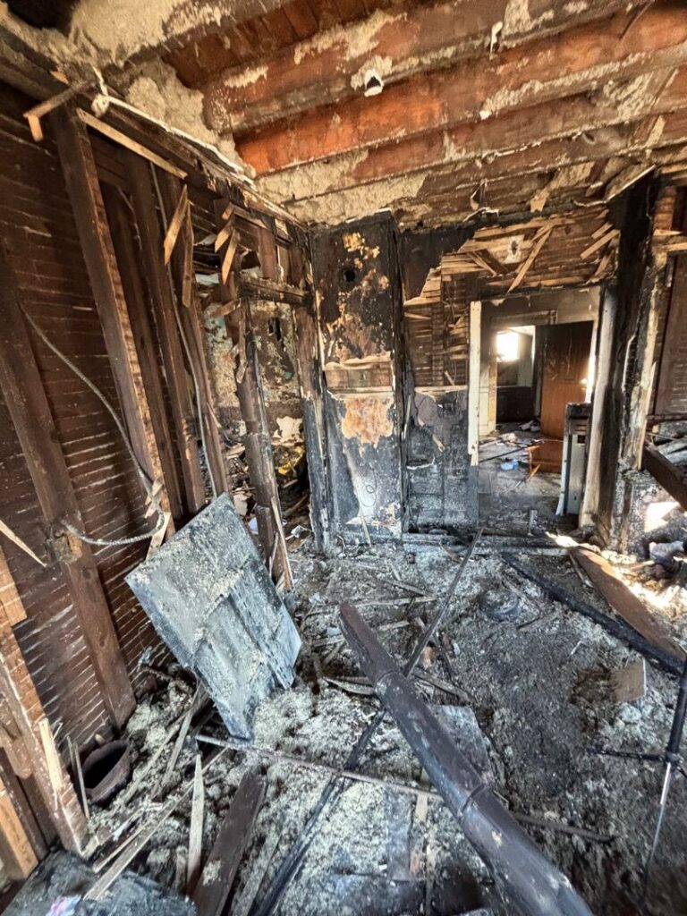An interior view showing severe structural damage from a house fire, with exposed wooden wall studs and floor debris.