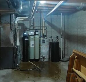 A utility room or basement area with a concrete floor showing a large, reflective puddle of standing water pooling around mechanical equipment. In the center, a water heater and several tall water filtration and softener tanks are lined up against a light gray brick wall. To the left is an HVAC unit with metal ductwork running along the ceiling, and a sump pump basin is visible in the right corner