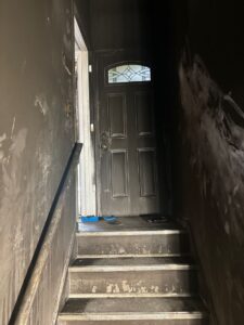 Dark narrow staircase showing severe smoke damage, heavy soot accumulation, and fire residue on the walls and steps leading up to a door.