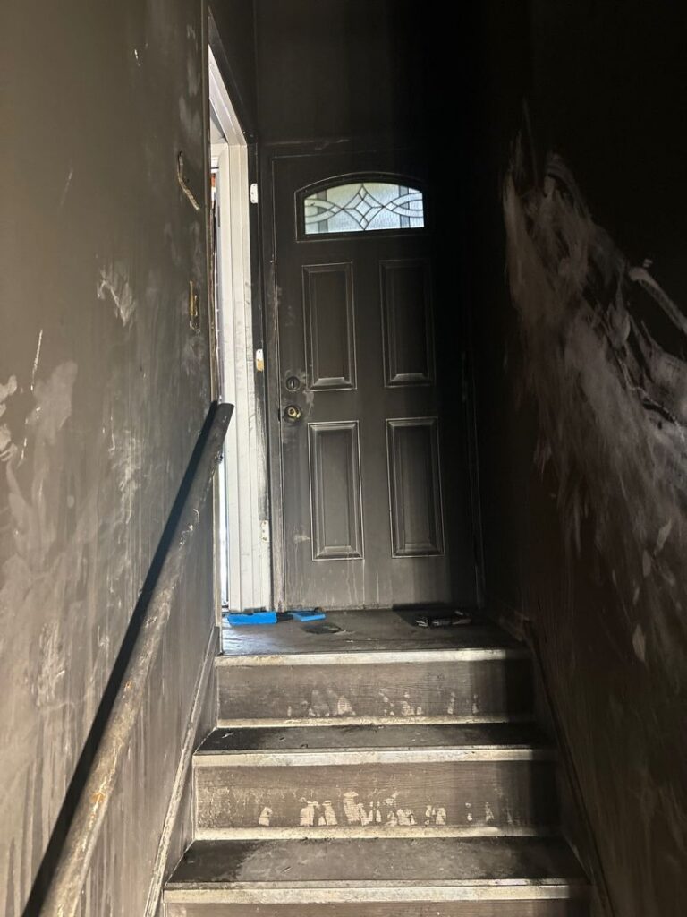 Dark narrow staircase showing severe smoke damage, heavy soot accumulation, and fire residue on the walls and steps leading up to a door.