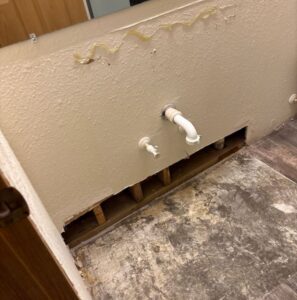 A close-up view of bathroom plumbing reveals evidence of water damage. Discoloration, peeling texture, and residual adhesive are visible on the painted wall surface. A white PVC pipe and another plumbing fixture emerge from the wall, with a lower section cut open to expose water-damaged subfloor and interior wall structure.