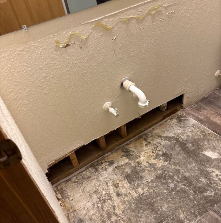 A close-up view of bathroom plumbing reveals evidence of water damage. Discoloration, peeling texture, and residual adhesive are visible on the painted wall surface. A white PVC pipe and another plumbing fixture emerge from the wall, with a lower section cut open to expose water-damaged subfloor and interior wall structure.