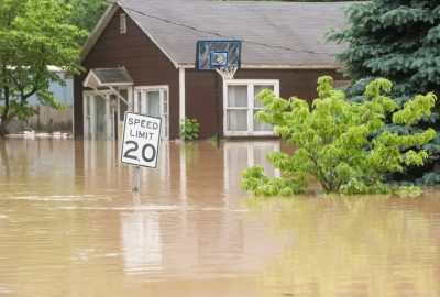A residential home partially submerged in brown floodwaters, with a speed limit sign and basketball hoop standing in the deep water, illustrating the dangers of flash flooding.
