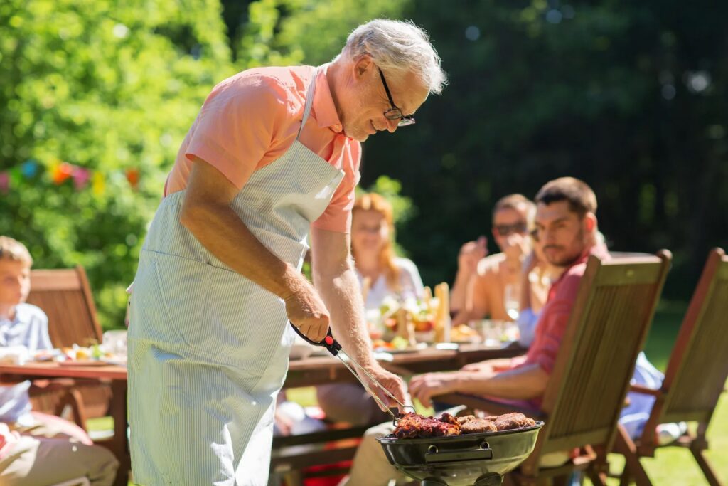 Older man in an apron grilling food on a sunny patio, with people at a table in the background having a meal together.