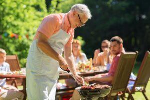 Older man in an apron grilling food on a sunny patio, with people at a table in the background having a meal together.
