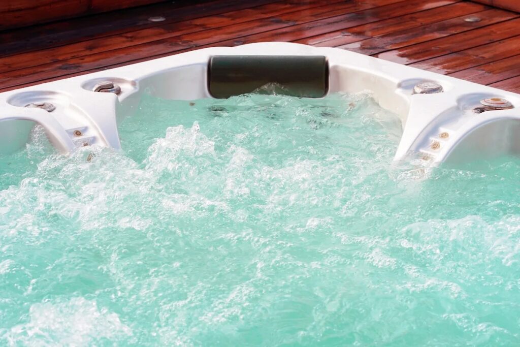 Close-up of a white hot tub with swirling, bubbly turquoise water, a dark headrest, and silver jets on a dark wood deck.