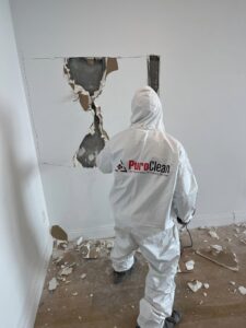A PuroClean technician in a white hazmat suit cuts a large hole in a white