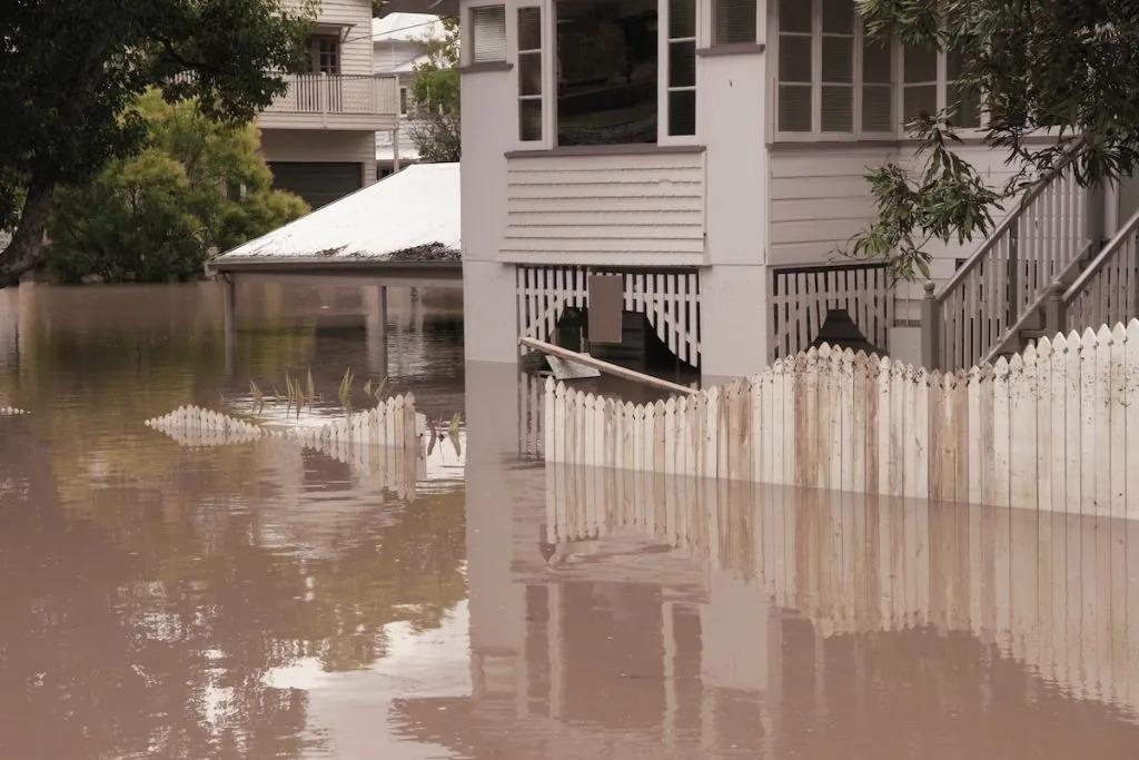 A residential house stands in deep, muddy floodwaters with a partially submerged picket fence.