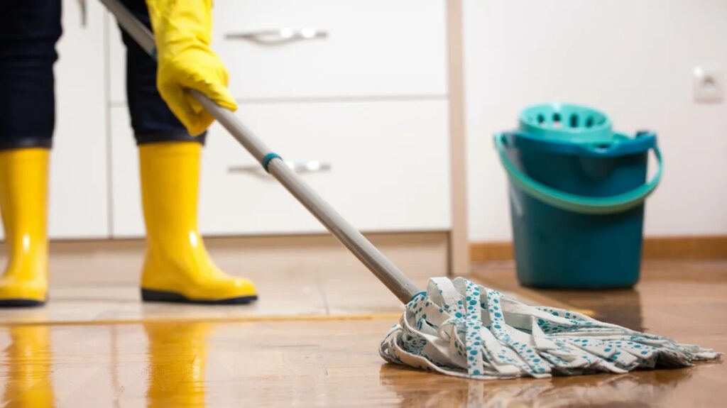 A person wearing yellow boots and gloves mops a shiny wooden floor next to a cleaning bucket.