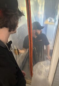 A technician holds a manometer to monitor air pressure while another technician observes through a plastic containment barrier.
