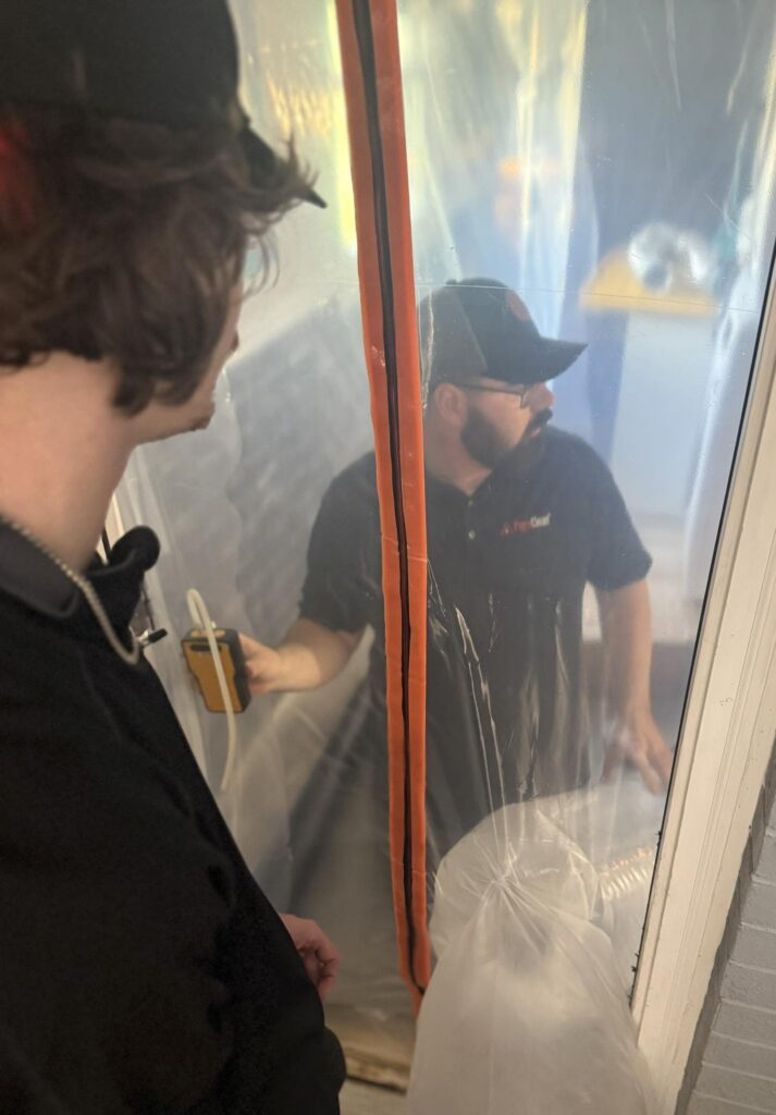 A technician holds a manometer to monitor air pressure while another technician observes through a plastic containment barrier.