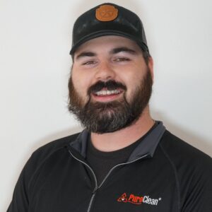 A bearded man wearing a black PuroClean shirt and hat smiles at the camera.