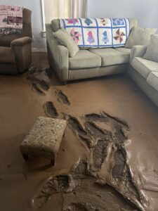 A living room floor is inundated with thick mud, featuring clear footprints and partially submerged furniture.