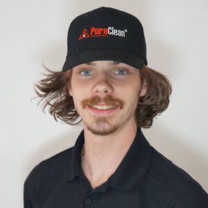 A smiling young man wearing a black PuroClean uniform cap and polo shirt looks forward.