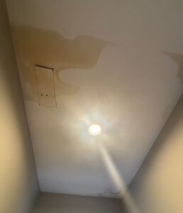 An upward-angled shot of a hallway ceiling showing a severe water stain spreading across the surface. The large, yellowish-brown water stain has distinct, irregular rings and covers a significant portion of the upper left side of the ceiling, surrounding a small rectangular access panel. A glowing, recessed circular light fixture in the center casts a bright glare downward, while smaller, fainter patches of water stain discoloration are visible along the upper right edges and corners of the ceiling.
