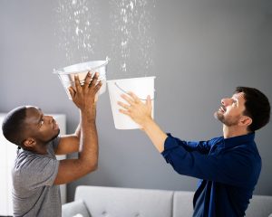 Two homeowners holding buckets to catch water leaking from the ceiling inside a living room.