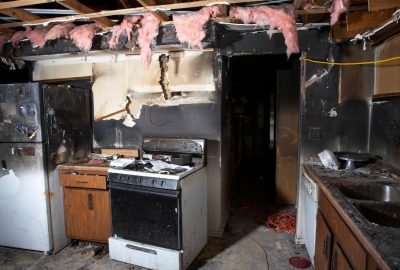 Fire-damaged kitchen with heavy smoke staining, exposed ceiling framing, and debris on the floor.