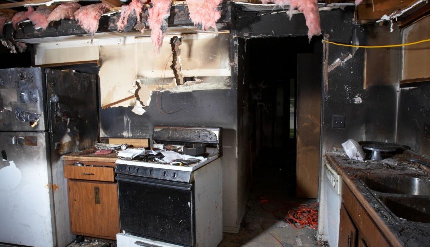 Fire-damaged kitchen with heavy smoke staining, exposed ceiling framing, and debris on the floor.