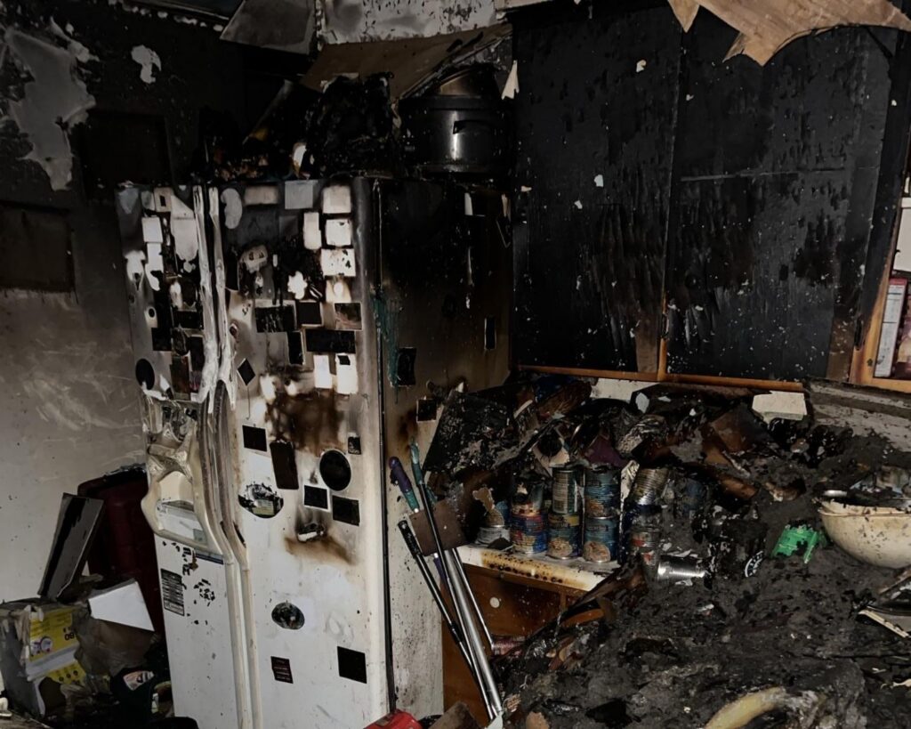 Fire-damaged kitchen with smoke-stained refrigerator, charred cabinets, and heavy soot and debris after an electrical fire.