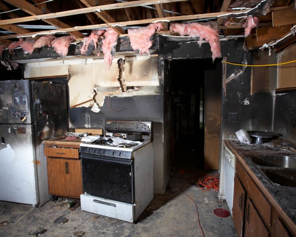 Fire-damaged kitchen with heavy smoke staining, exposed ceiling framing, and debris on the floor.