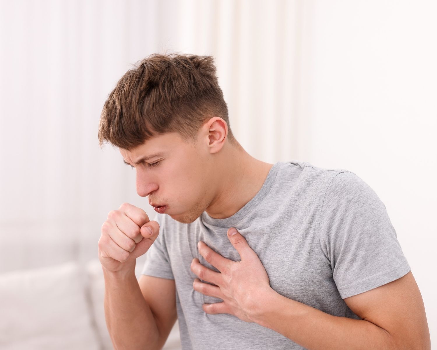 Young man coughing into his fist and holding his chest while standing inside a home