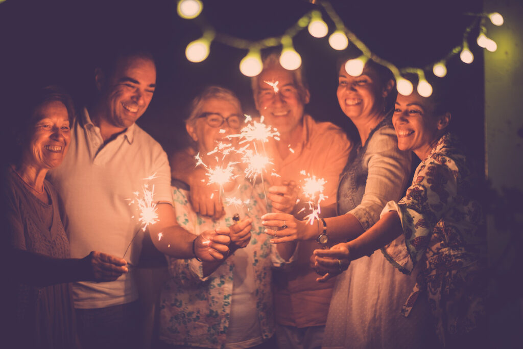 A group of six people joyfully holding sparklers, celebrating together under string lights on New Year's Eve.