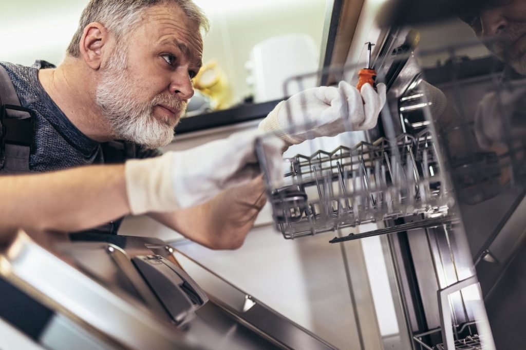 A figure with a screwdrver ensuring mounting screws on a the top inside of a dishwasher are tight so the door seal is held firmly in place and does not cause a dishwasher leak