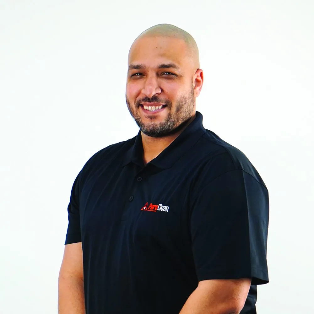Smiling man with a bald head wearing a black polo shirt, standing against a plain white background.