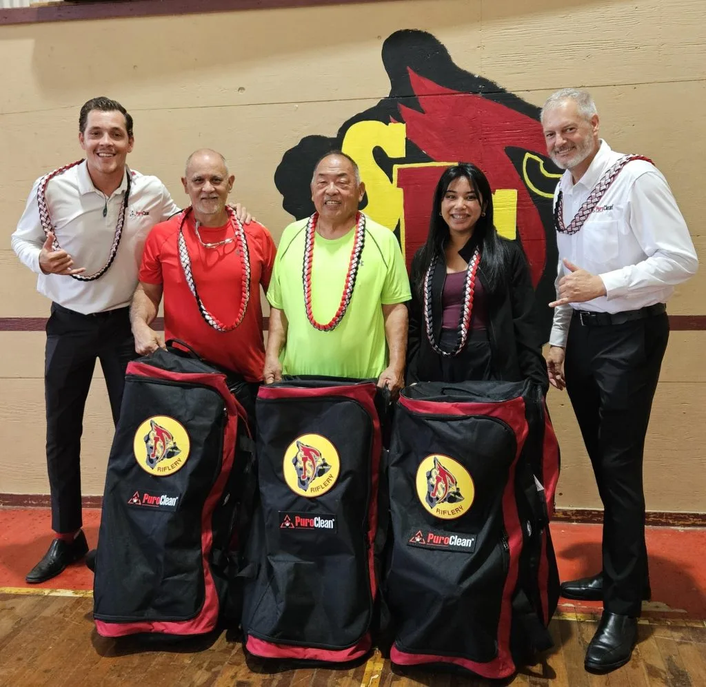 Five people in colorful attire stand with large bags, smiling in front of a wall featuring a cardinal logo.